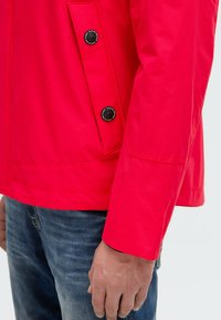 Veste rouge à la texture lisse, avec deux boutons noirs sur le côté gauche et un design légèrement court. Portée sur un jean.