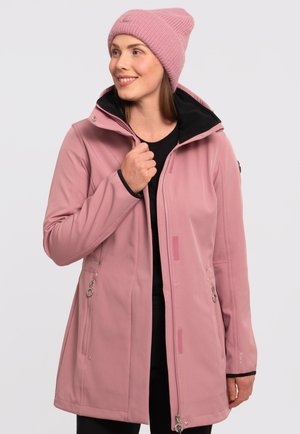Veste softshell rose clair avec un intérieur noir, poches latérales zippées et col montant. Portée avec un bonnet en tricot rose assorti.