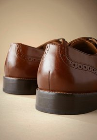 Chaussures habillées en cuir marron avec une finition lisse, des détails de brogue et un talon noir solide, montrées de l'arrière en gros plan.