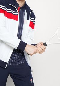 White jacket with red and navy stripes, navy striped polo, and navy shorts. Holding a tennis racket, showcasing sporty design and colors.
