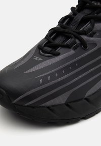 Baskets de sport noires et grises avec une surface texturée, des panneaux en maille, des rayures distinctes et un col rembourré ; elles présentent des lacets noirs et un détail de logo.