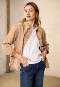 Naine kannab beežikat jopet valge särgi ja siniste teksade üle, seisab siseruumides käed taskus, vaatab enesekindlalt kaamerasse.