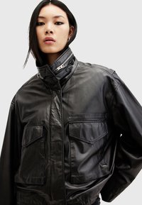 Black leather jacket featuring a high collar, two chest pockets, and silver zipper detailing. Smooth texture with a relaxed fit.