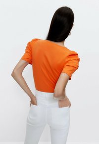 Oranžový fitted top s balónovými rukávy, zkombinovaný s bílými vysokými džínami s velkými zadními kapsami. Hladká bavlněná textura.