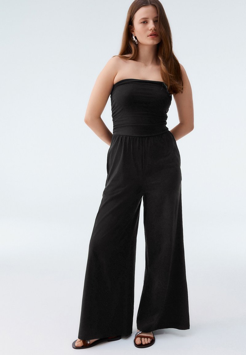 Black strapless jumpsuit with a fitted bodice and wide-leg trousers. Smooth fabric with a relaxed fit. Features pockets and minimal detail.
