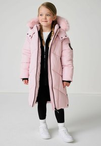 Cappotto puffo rosa con cappuccio foderato in pelliccia, chiusure a zip e bottoni a pressione, caratterizzato da un design trapuntato. Indossato sopra un outfit nero e sneakers bianche.