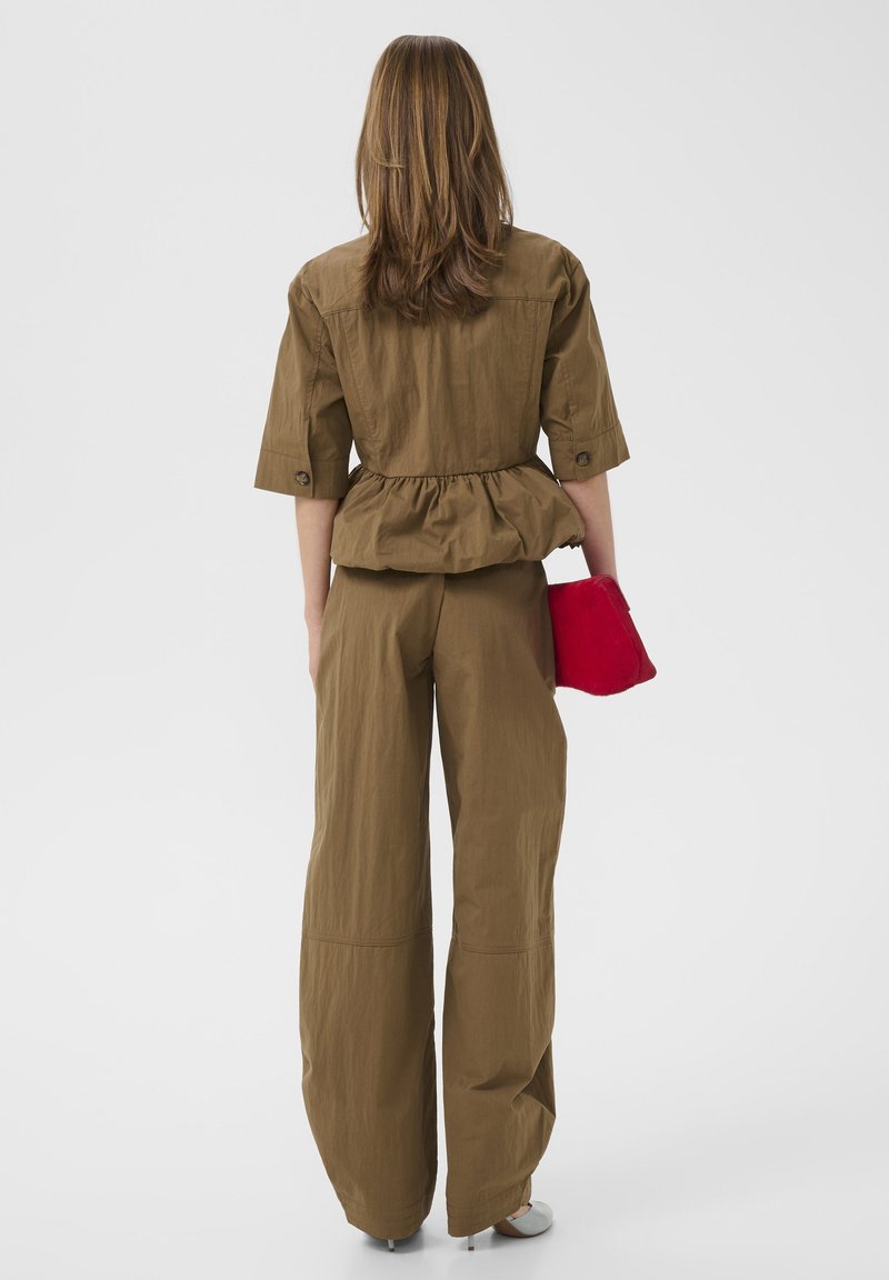 Vrouw in een bruin jasje met geplooide taille en wijde pijpen broek, die een felrode clutch vasthoudt en voor een effen witte achtergrond staat.