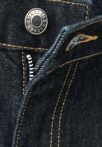 Tela vaquera con costura amarilla que presenta un botón metálico y una cremallera plateada parcialmente abierta en la cintura de unos vaqueros.