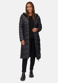 Zwarte pufferjas met capuchon, voorzien van een chevron-quiltontwerp, zijzakken en een middenlange zoom. Gedragen over een zwart topje en een broek.