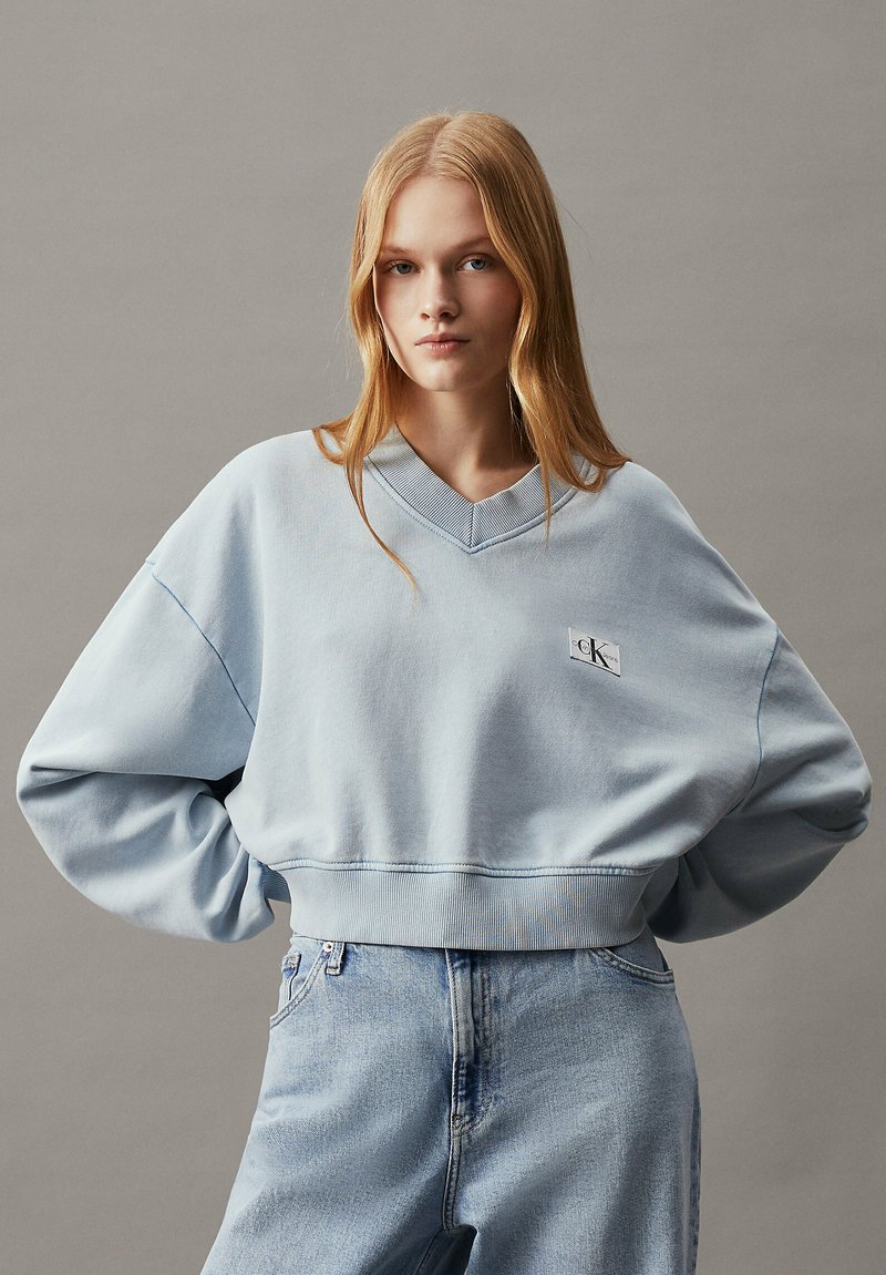 Felpa azzurra oversize con scollo a V, polsini e orlo a costine. Presenta una piccola toppa con logo sul davanti. Indossata con jeans azzurri.