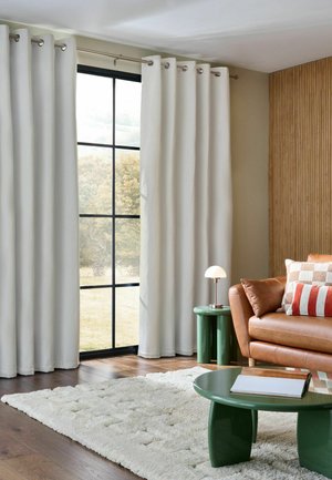 Wohnzimmer mit großem Fenster und weißen Vorhängen, braunes Ledersofa mit gemusterten Kissen, grüne Beistell- und Couchtische, weißer Teppich auf Holzboden.