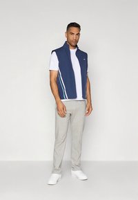 Tamsiai mėlyna liemenė be rankovių su baltomis ir šviesiai mėlynomis juostelėmis, dėvima ant baltos palaidinės, derinama su pilkais sportinėmis kelnėmis ir baltais sportbačiais.