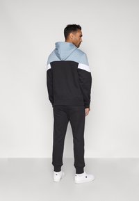 Homem em pé, vestindo calças de treino pretas, uma camisola com capuz em blocos de cor nas cores preta, azul claro e branca, e ténis brancos com logótipos pretos.