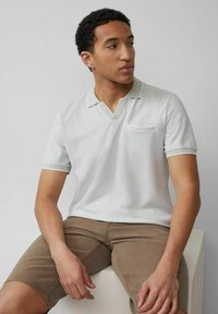 Hellgraues Polohemd mit kurzem Kragen und Tasche, mit kontrastierendem Besatz am Kragen und an den Ärmeln. Das Modell trägt braune Shorts.