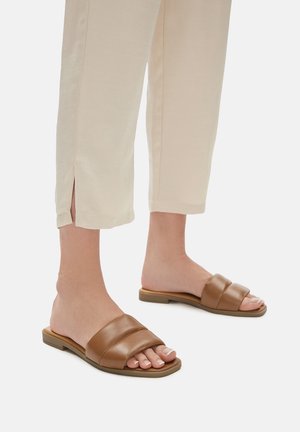 Hnedé kožené šľapky s dvoma širokými popruhmi, plochou podrážkou a hladkým povrchom, pričom sú skombinované so svetlými skrátenými nohavicami.