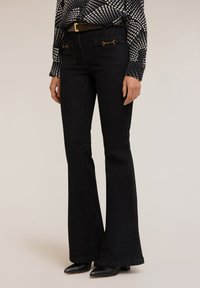 Jeans negros de campana con cintura alta, que presentan acentos de hardware en tono dorado en los bolsillos traseros, combinados con una blusa de estampado blanco y negro.