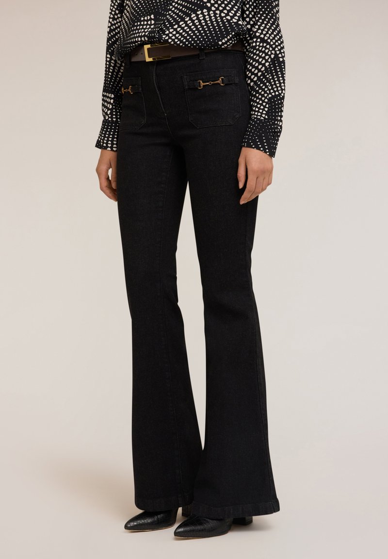 Jeans negros de campana con cintura alta, que presentan acentos de hardware en tono dorado en los bolsillos traseros, combinados con una blusa de estampado blanco y negro.