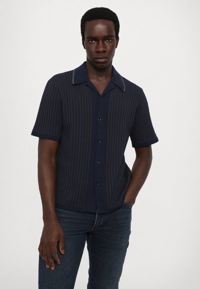 Kortermet skjorte i marineblått med vertikale striper, knappefront, skjortekrage, kontrastkanting og subtilt teksturert mønster. Brukt med mørke jeans.