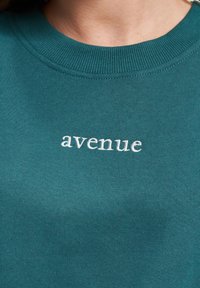 Sjøgrønn hettegenser med ribbestrikket halsringning og brodert "avenue" tekst i hvitt, laget av et mykt stoff med glatt tekstur.