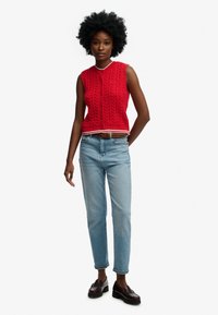 Kvinde med afro-hår iført en rød ærmeløs knaptrøje, lyseblå jeans med brunt bælte og mørkebrune loafers, står med den ene hånd bag ryggen.