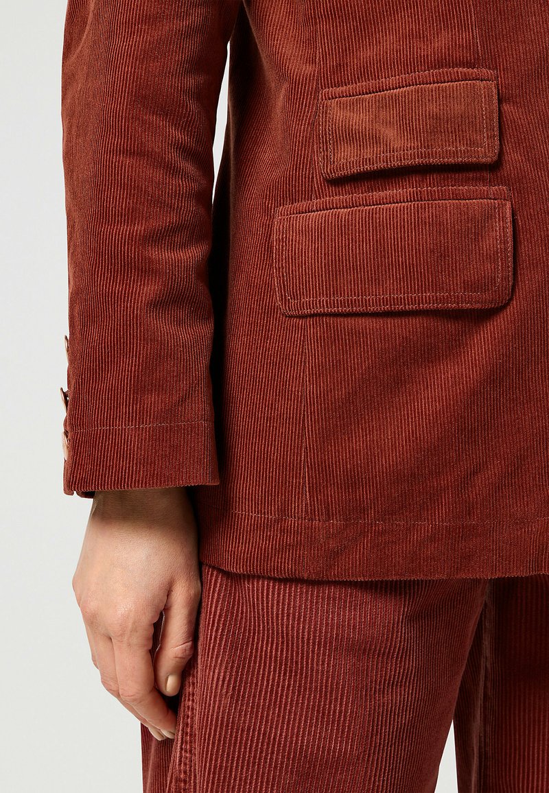 Roodkleurig corduroy-pak met verticale ribbels, voorzien van een drieknoopsluiting en twee opgestikte zakken aan de achterkant. Gladde textuur.