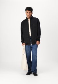 Black quilted jacket with high collar, zip closure, and a logo on the chest. Paired with light sweater, blue denim, and a beige tote bag.