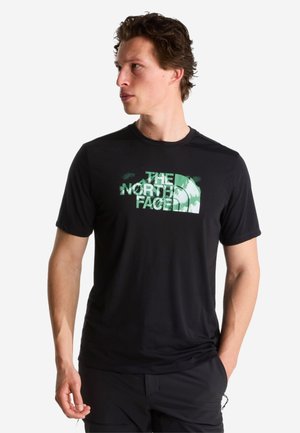 Mand iført en sort The North Face T-shirt med grøn logo, ser til venstre med den ene hånd i lommen og neutral baggrund.