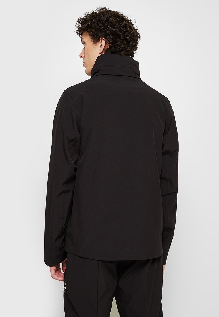 Giacca nera con colletto alto, caratterizzata da una texture liscia e un design pulito e minimale. Il modello è visto di spalle, mettendo in evidenza la vestibilità.