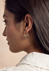 Goldfarbene Creolen mit silbernen geometrischen Anhängern, die ein strukturiertes, offenes Design aufweisen. Das Modell hat dunkle Haare und trägt ein weißes Oberteil mit Kragen.