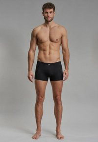 Black boxer shorts made from stretch fabric, featuring a soft waistband. The model stands barefoot against a neutral gray background.