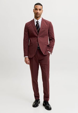 Homme debout portant un costume bordeaux avec une chemise blanche, une cravate noire rayée et des chaussures noires sur un fond clair uni.