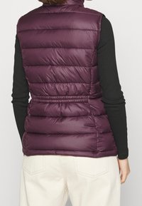 Prošívaná vesta v tmavě fialové barvě s vysokým límcem, obsahující horizontální prošívané sekce a nastavitelný šňůrkový pas. Klasický střih bez kapes.