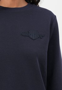 Persoană purtând un top bleumarin cu mânecă lungă, având un logo brodat închis pe o patcă pe partea stângă a pieptului, pe care scrie "GANT."