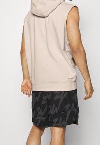 Sleeveless beige hoodie with a hood, relaxed fit, paired with black shorts featuring a bold, dark gray lightning bolt pattern.