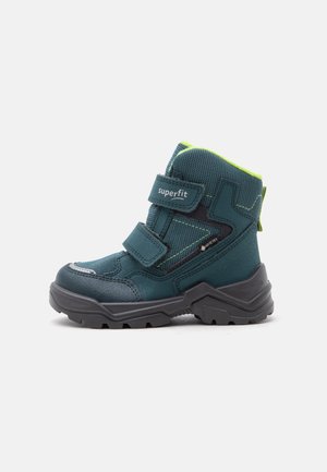 Otroški pohodniški čevelj temno modro-zelene in črne barve z dvema ježkoma, oznaka "superfit", Gore-Tex tkanina in debel robusten podplat na belem ozadju.