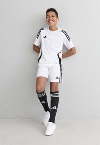 Jersey y pantalones cortos blancos de Adidas con acentos negros, combinados con calcetas negras hasta la rodilla. Las características incluyen tres franjas negras y un diseño deportivo.