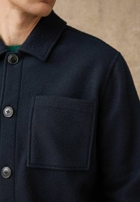 Granatowy wełniany płaszcz z kołnierzem, czarnym zapięciem na guziki oraz jedną prostokątną kieszenią na lewej piersi. Miękka tekstura.