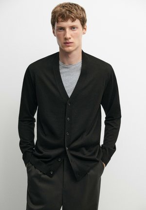 Junger Mann mit heller Haut und kurzen roten Haaren, der einen schwarzen, geknöpften Cardigan über einem grauen T-Shirt trägt und die Hände in den Taschen hat.