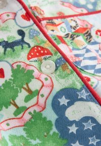 Monivärinen kangas, jossa on leikkisiä kuvioita puista, sienistä ja eläimistä. Punainen reunus ja valkoinen nappi täydentävät muotoilua.
