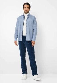 Camicia blu a bottoni con taschino sul petto indossata sopra un dolcevita bianco, abbinata a pantaloni in velluto a coste blu navy e sneakers bianche. Sfondo semplice.