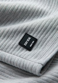 Tessuto grigio a righe con una superficie testurizzata e un'etichetta rettangolare nera che recita "STAY REAL SINCE 1985" attaccata ad esso.