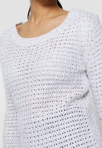 Pull en maille blanche avec un motif ouvert et texturé, un large col rond et des manches longues. Le tissu est léger et respirant.