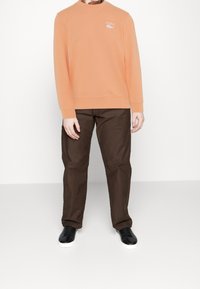 Sweater arancione con collo rotondo a costine e logo Lacoste bianco, abbinato a pantaloni marrone scuro e sneakers nere con suole bianche.