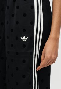 Μαύρο παντελόνι με ανάγλυφα πουά, λευκό λογότυπο Adidas trefoil και τρεις λευκές κάθετες ρίγες δίπλα σε ένα ελαφρώς μαυρισμένο χέρι που ακουμπά κοντά στο μηρό.