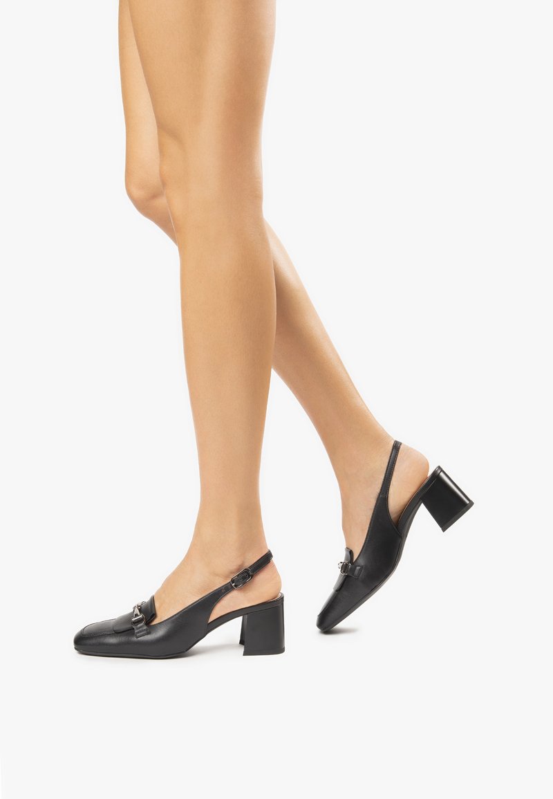 Tacchi slingback in pelle nera con punta quadrata, tacco a blocco e decorazioni hardware nella parte anteriore. Texture liscia e design minimale.
