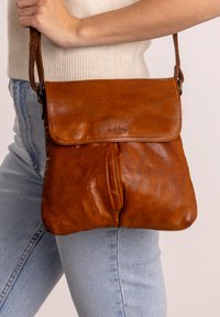Brun læder crossbody taske med klap holdt af en person iført lyseblå jeans og en cremefarvet strikket top.