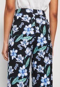 Pantalons à imprimé floral avec un fond noir et des fleurs blanches et bleues, taille haute, et tissu texturé avec des coutures visibles.