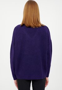 Personne aux cheveux roux raides, mi-longs, portant un pull ample de couleur violet foncé et un pantalon noir, vue de dos sur un fond blanc.