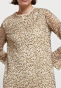 Béžový top s priesvitnými dlhými rukávmi, zdobený zlatými a hnedými flitrami v rozptýlenom vzore. Okrúhly výstrih s jemným dokončením.