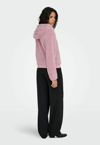 Nepbont hoodie in zachtroze, met een capuchon en geribbelde manchetten. Gecombineerd met wijde zwarte broek en laarzen met hak. Zicht vanaf de zijkant.